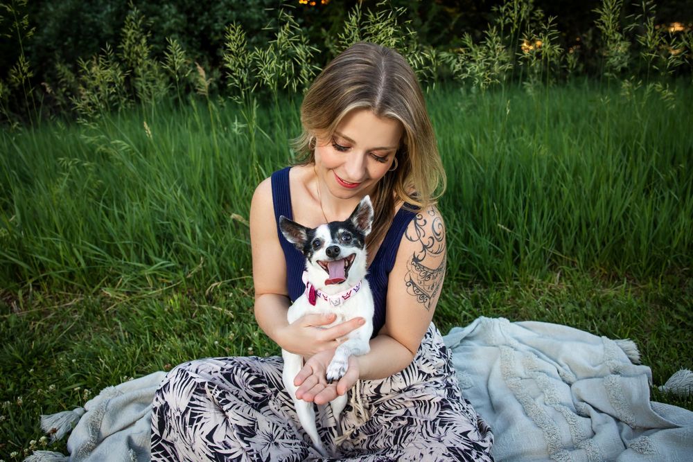 Woman with tattoos sitting on blanket holding small dog in green field.
