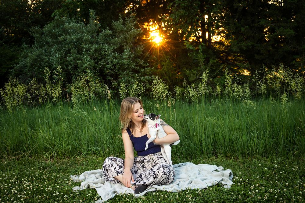 Woman sitting on blanket with small dog in green field at golden hour.