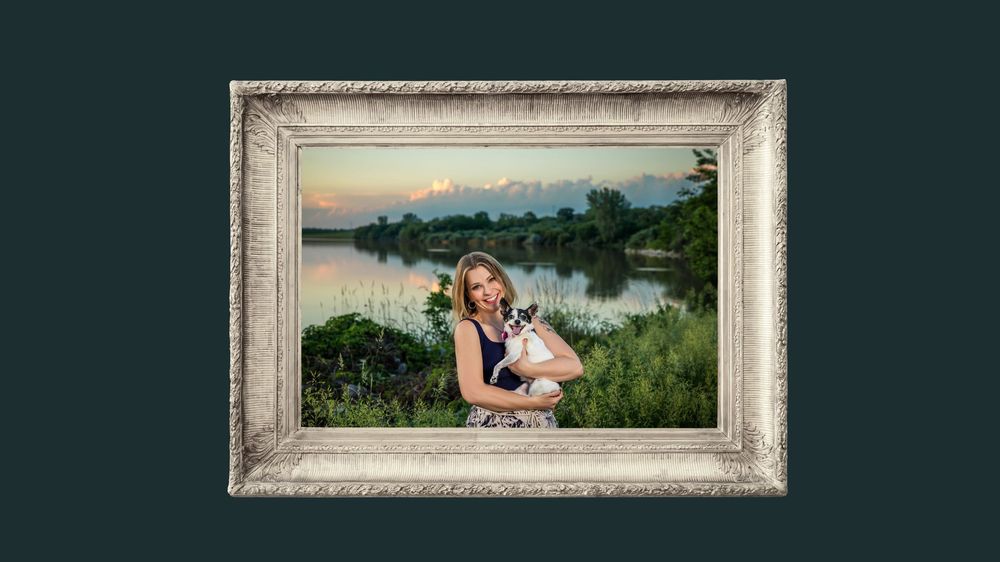 Smiling woman holding small dog in decorative picture frame with lake backdrop.
