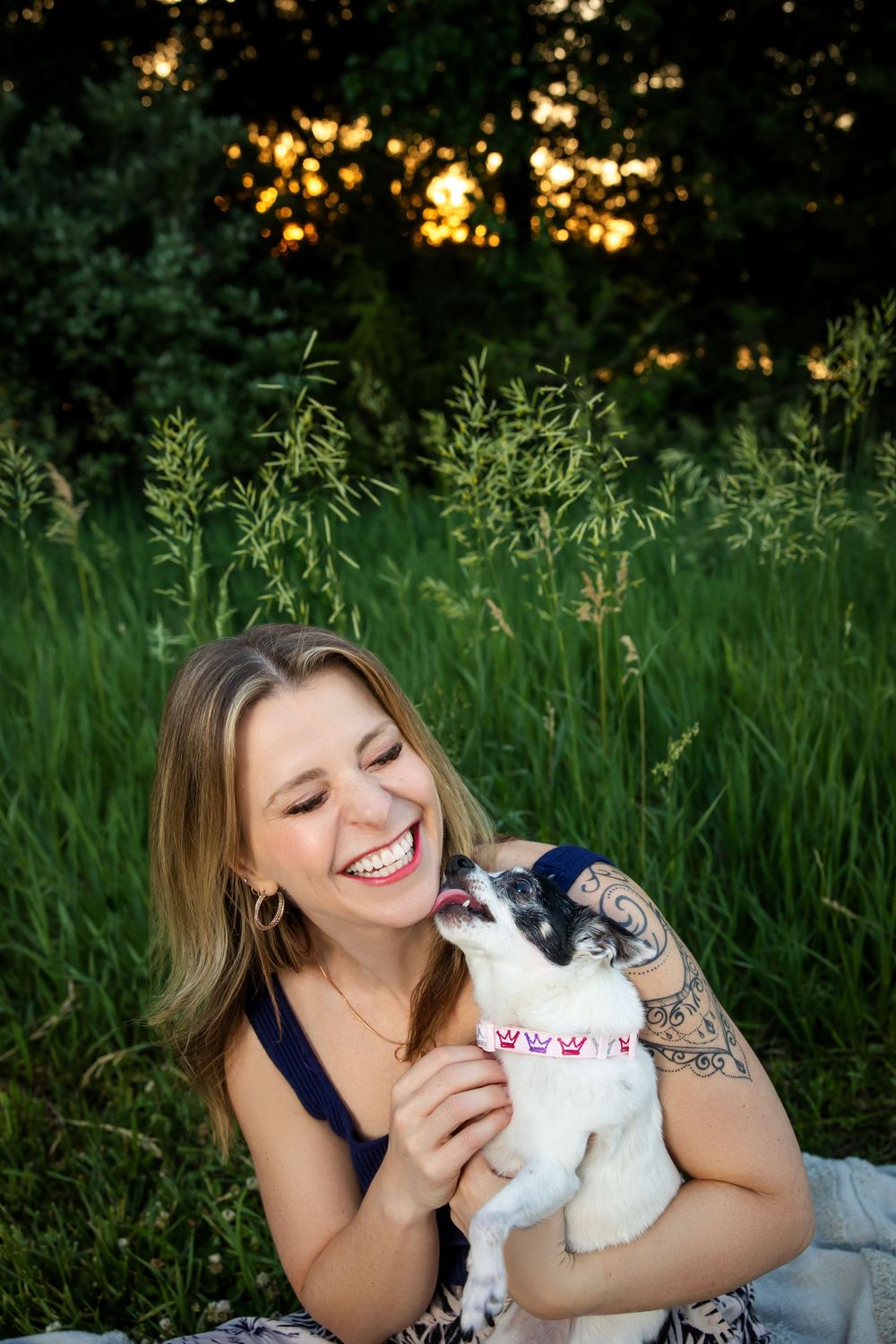 Laughing woman with tattoos holding small dog licking her face outdoors.