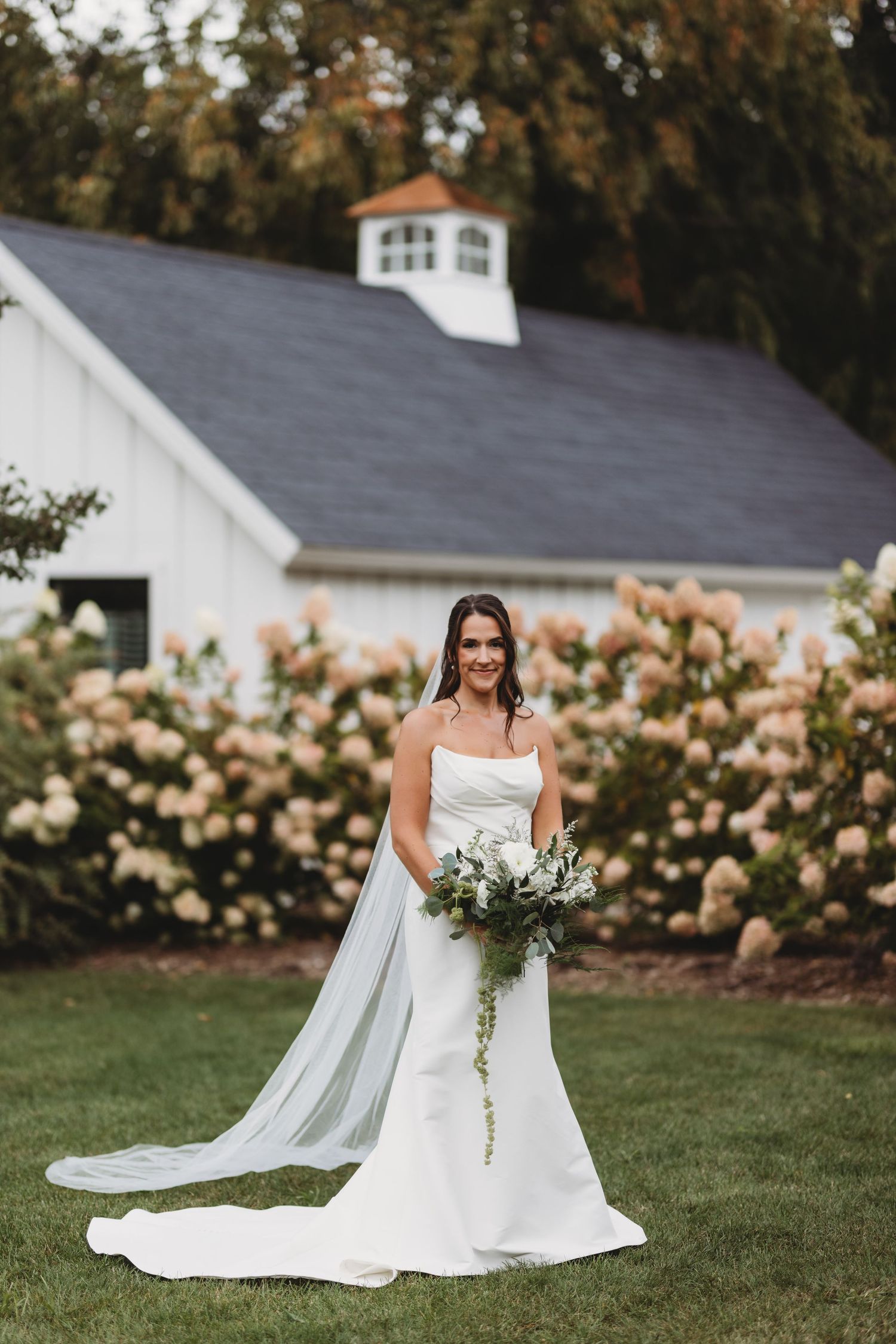 Bride in white strapless gown holding greenery bouquet stands on lawn before white barn and blooming hydrangeas.