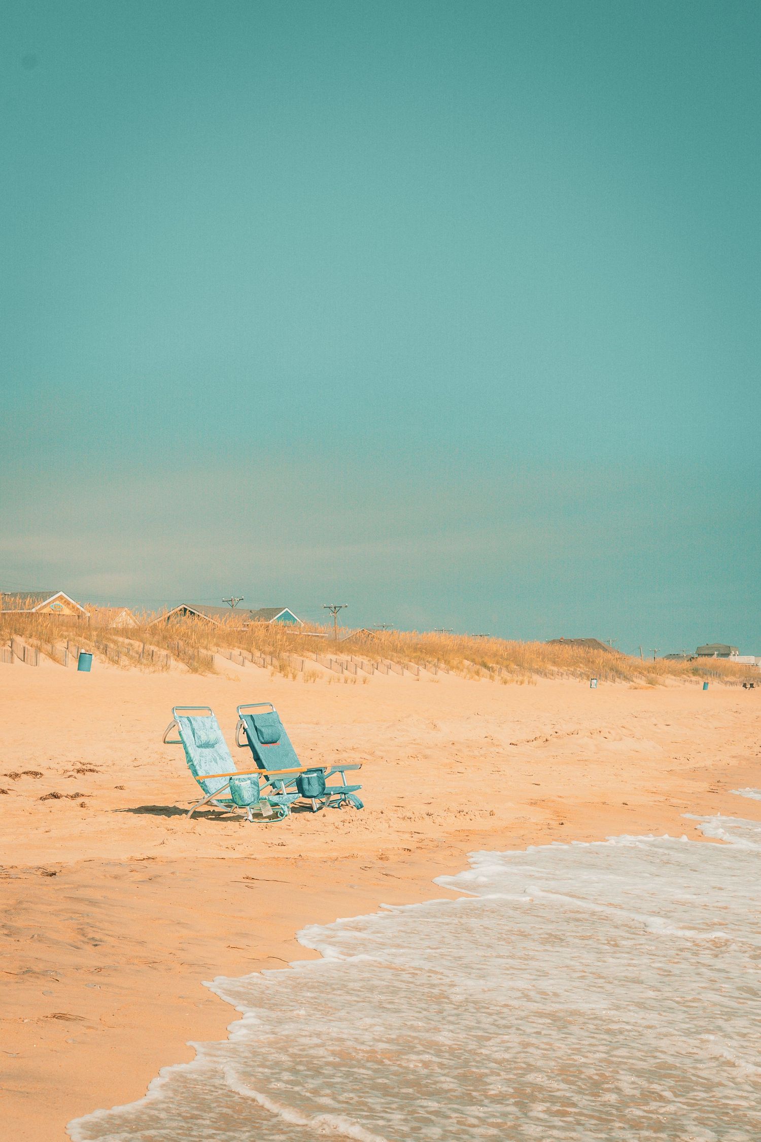 Two turquoise beach chairs sit on sandy shore with gentle waves and dune grass under a clear teal sky.