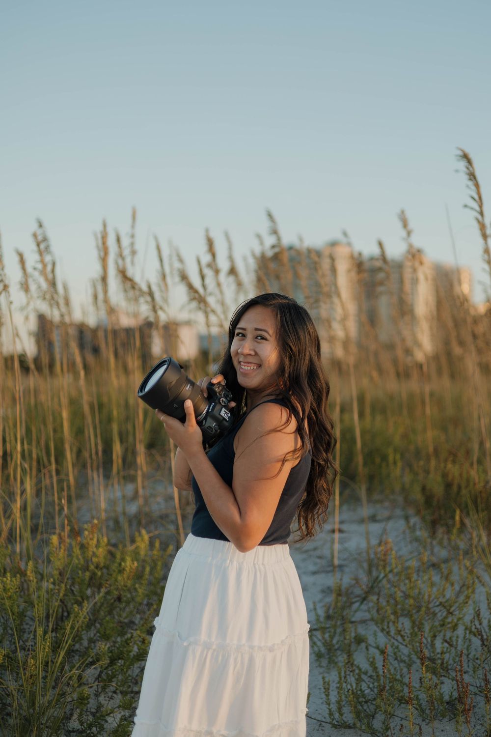 Young woman smiling and holding a camera on a sandy beach with sea oats at golden hour.