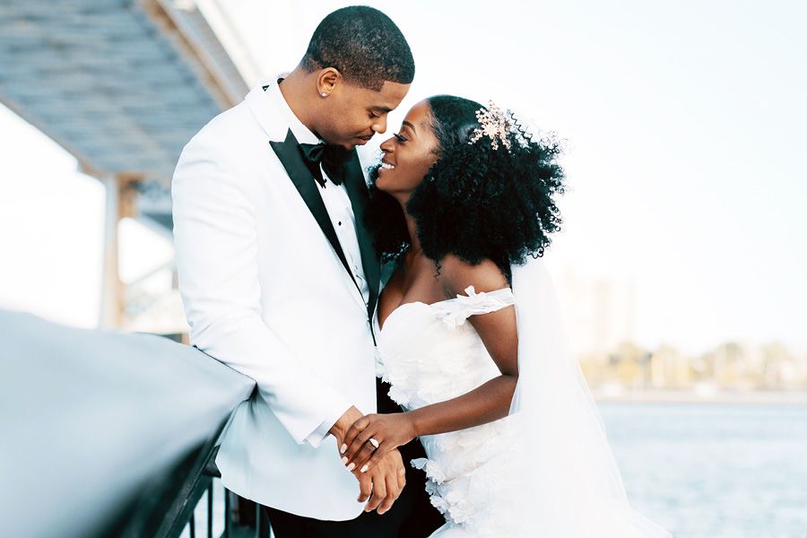 A Black couple shares a romantic moment in wedding attire, groom in white tuxedo, bride in off-shoulder gown, near waterfront.