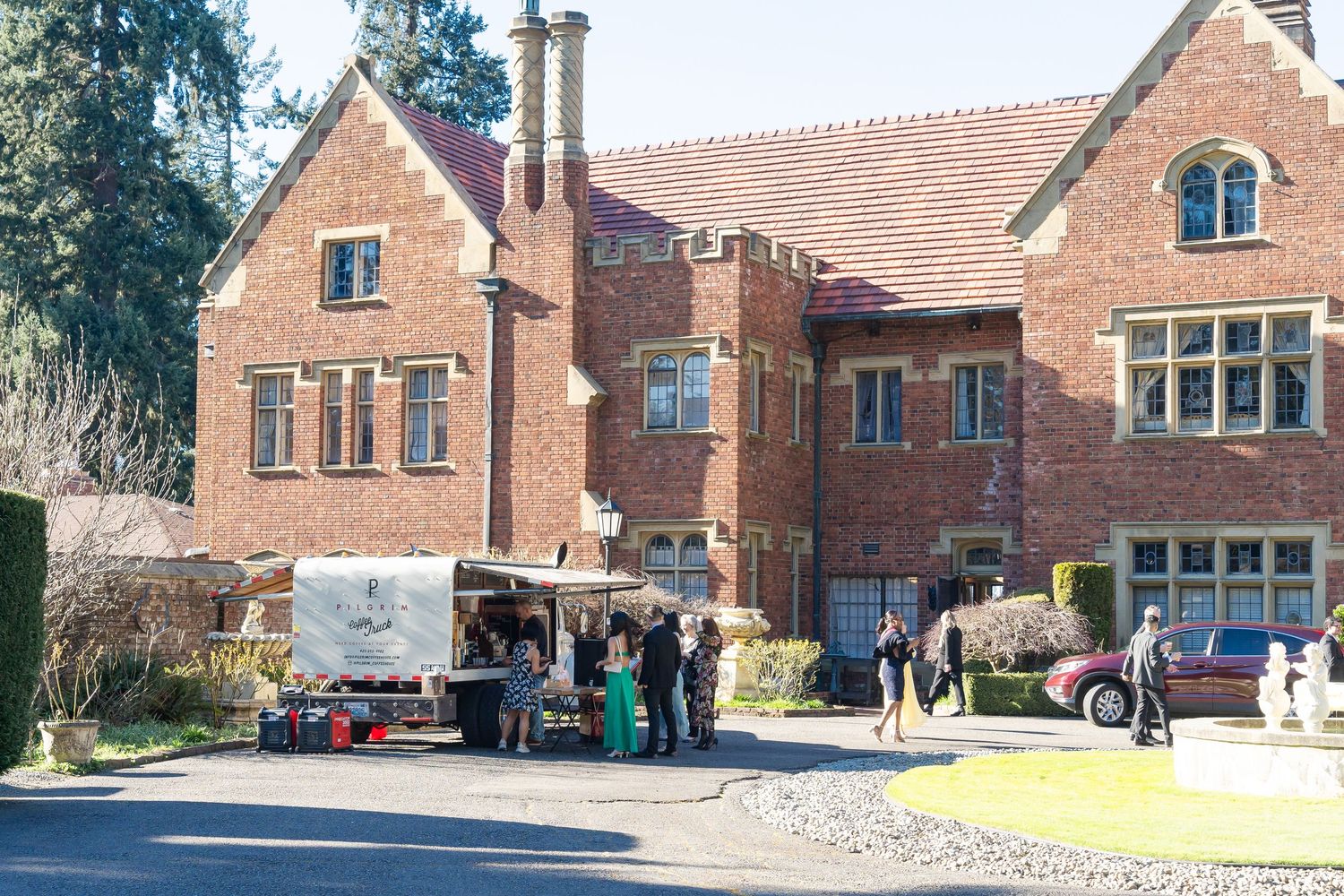 Elegant brick manor house with guests and catering truck outside during a formal event on a sunny day.
