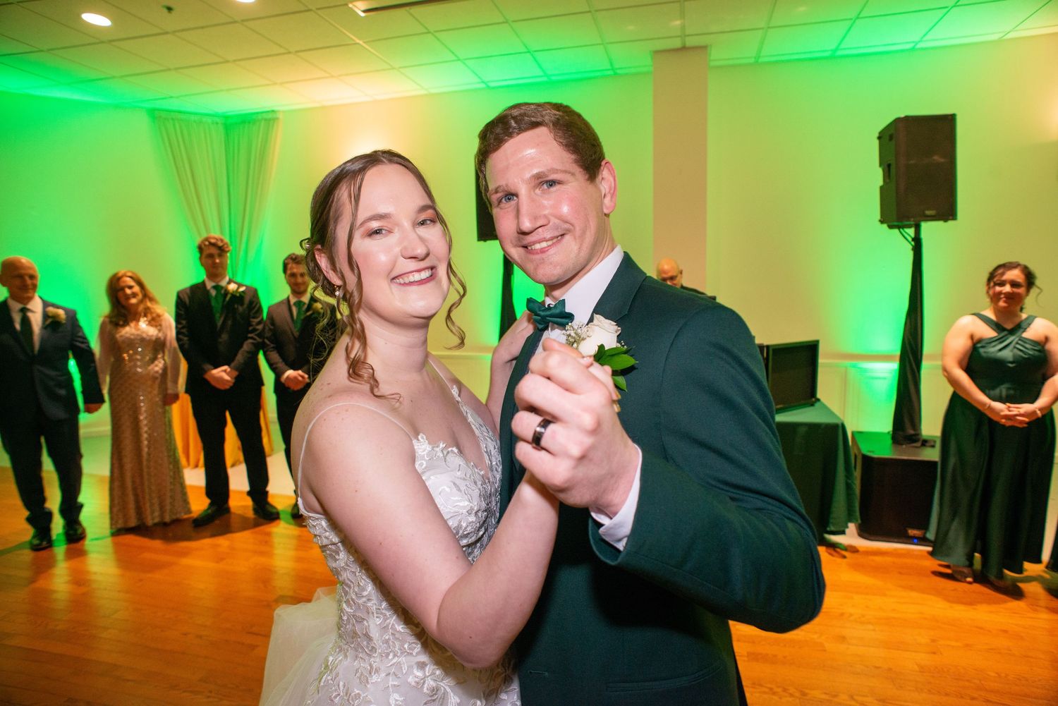 Bride and groom share first dance at wedding reception with green uplighting and wedding party in background.