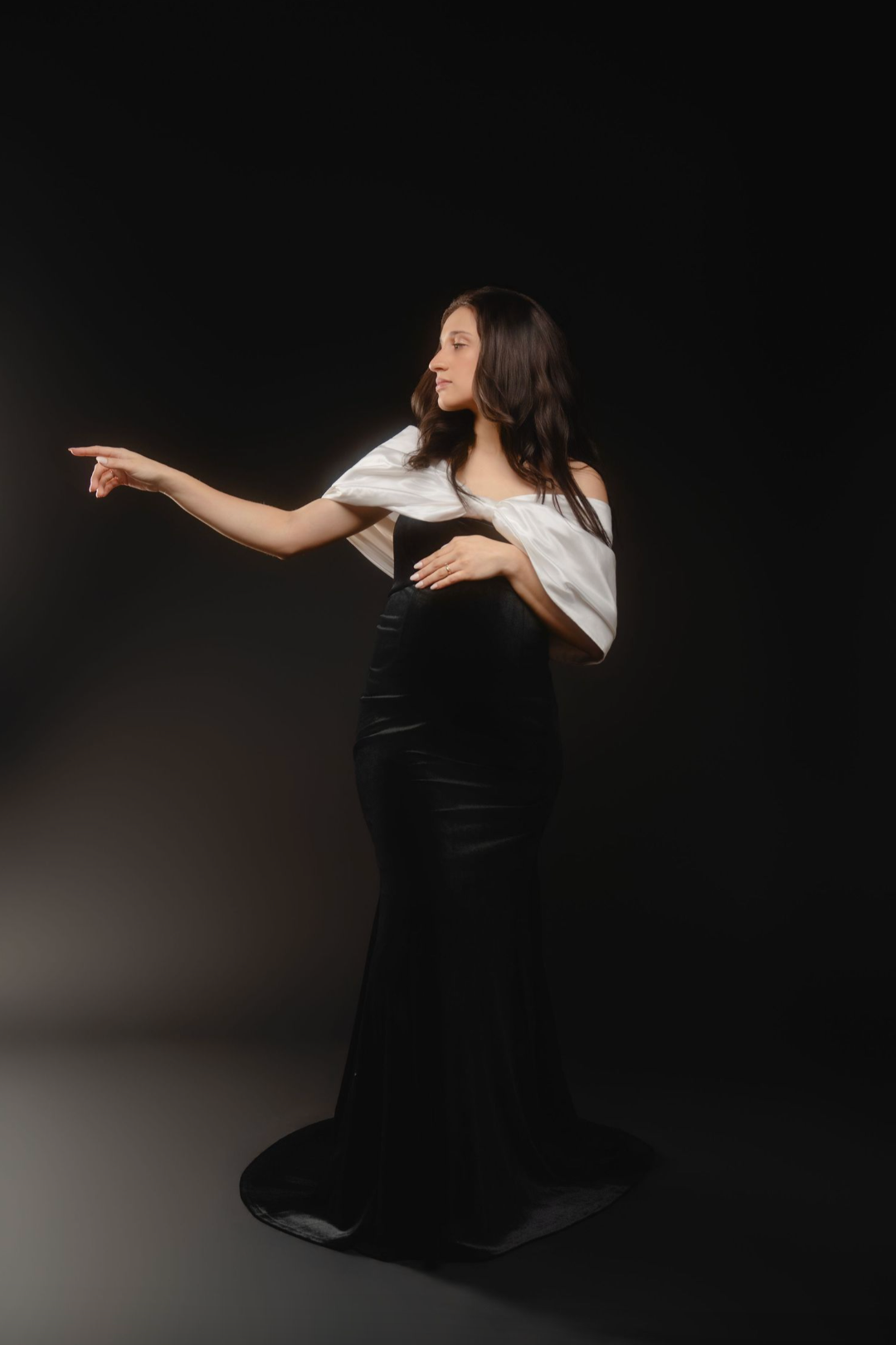 Elegant woman in black skirt and white off-shoulder top pointing sideways on dark background.