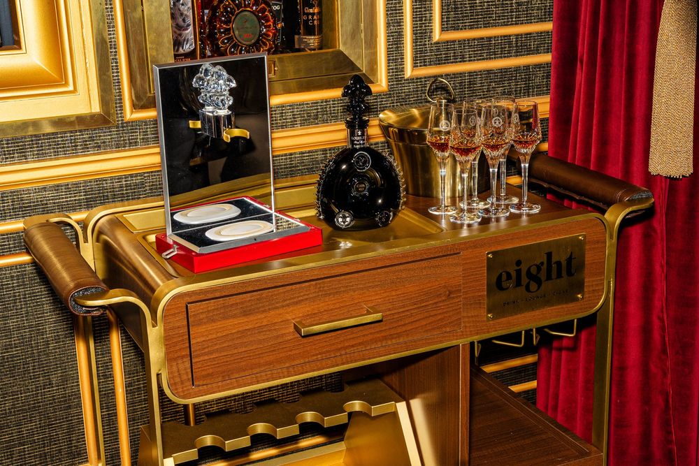 Luxury gold bar cart with cognac decanter, champagne flutes, and elegant Eight branding in opulent interior.
