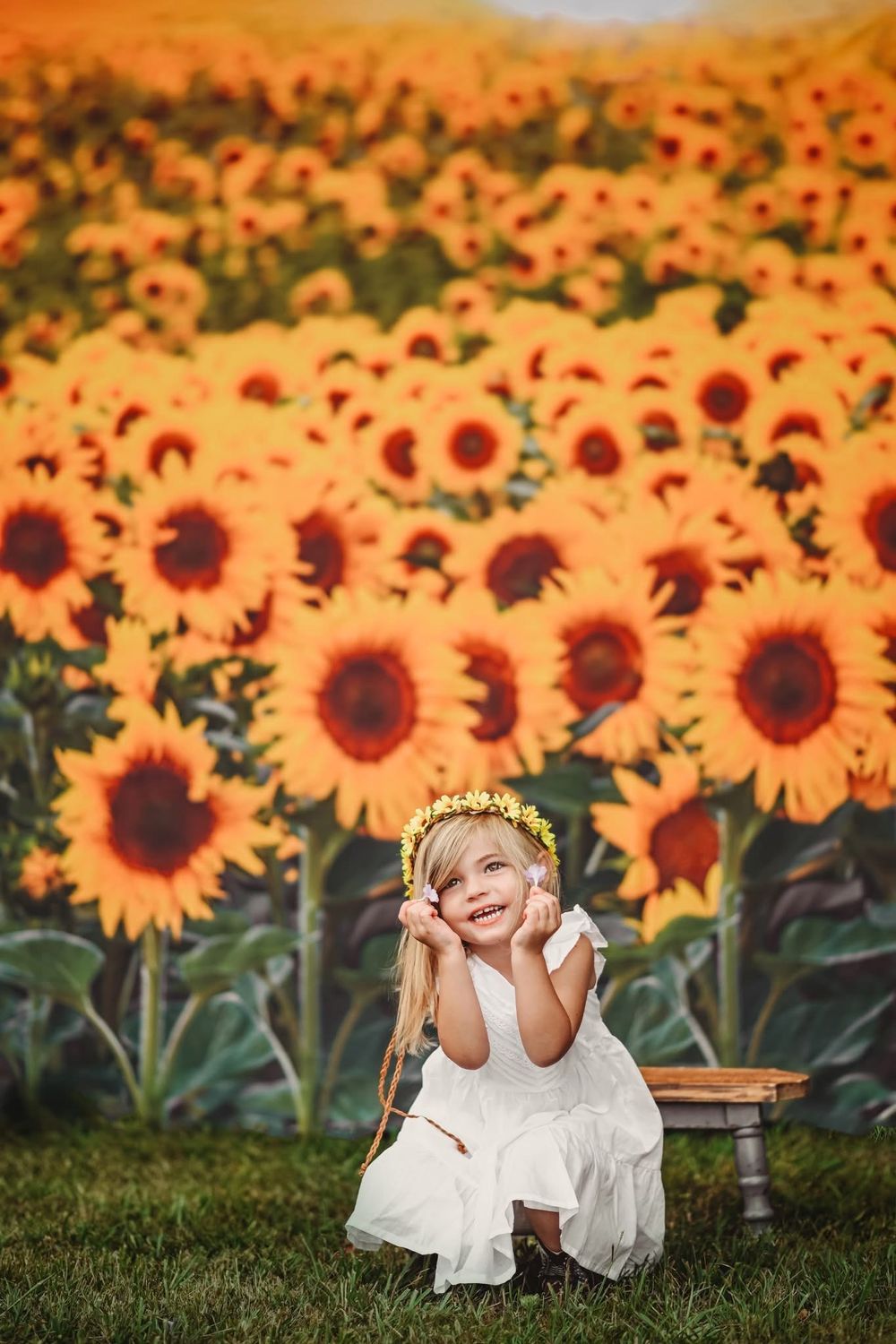 Young girl in white dress and floral crown sits smiling before a vibrant sunflower field backdrop.