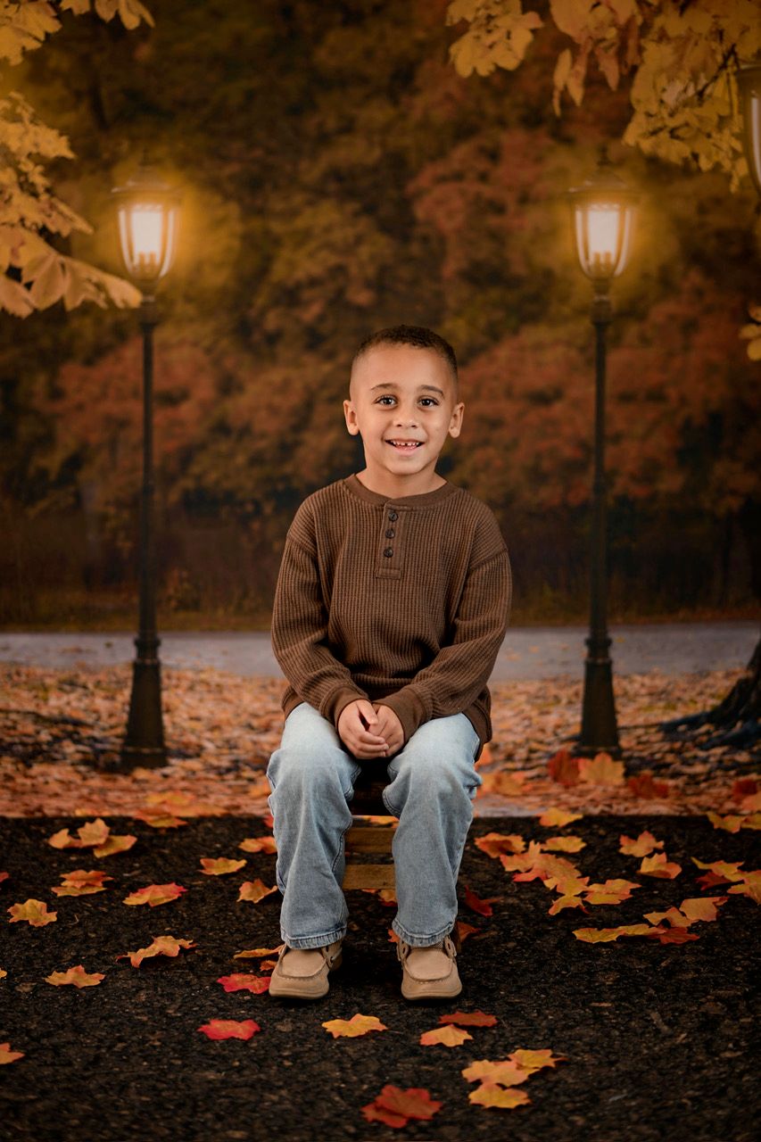 A smiling young boy sits on a bench surrounded by autumn leaves, framed by glowing street lamps and colorful fall foliage.