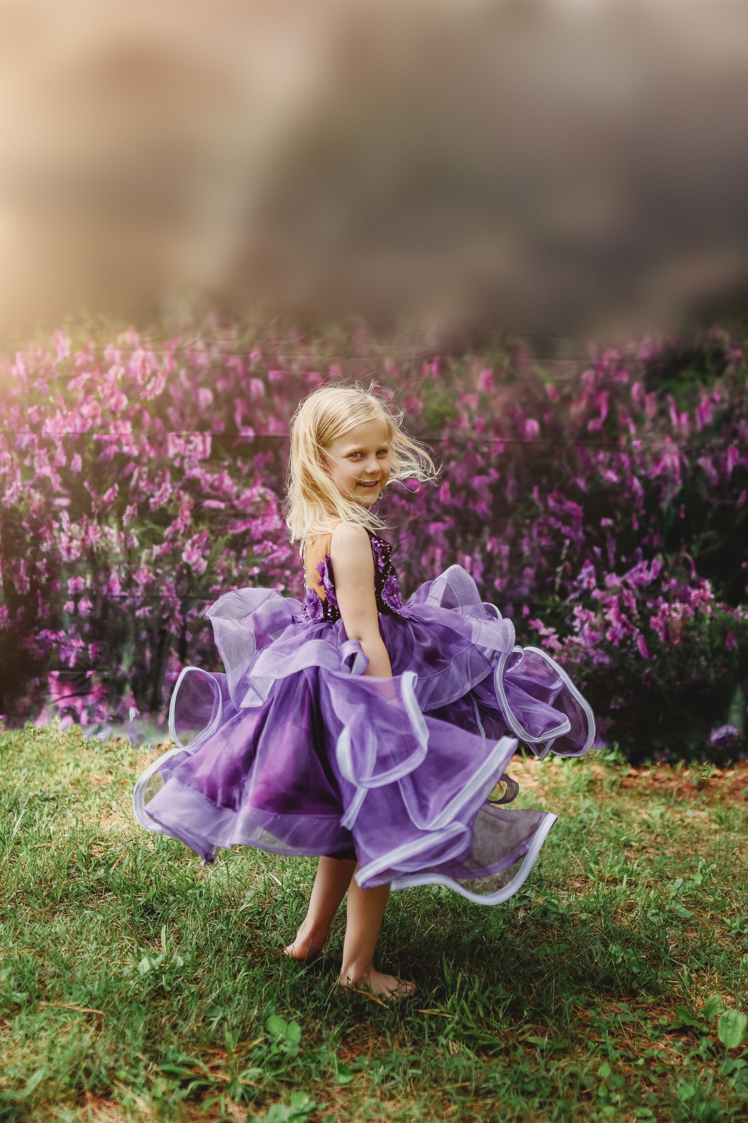 Young blonde girl twirling in purple ruffle dress outdoors among pink flowers.