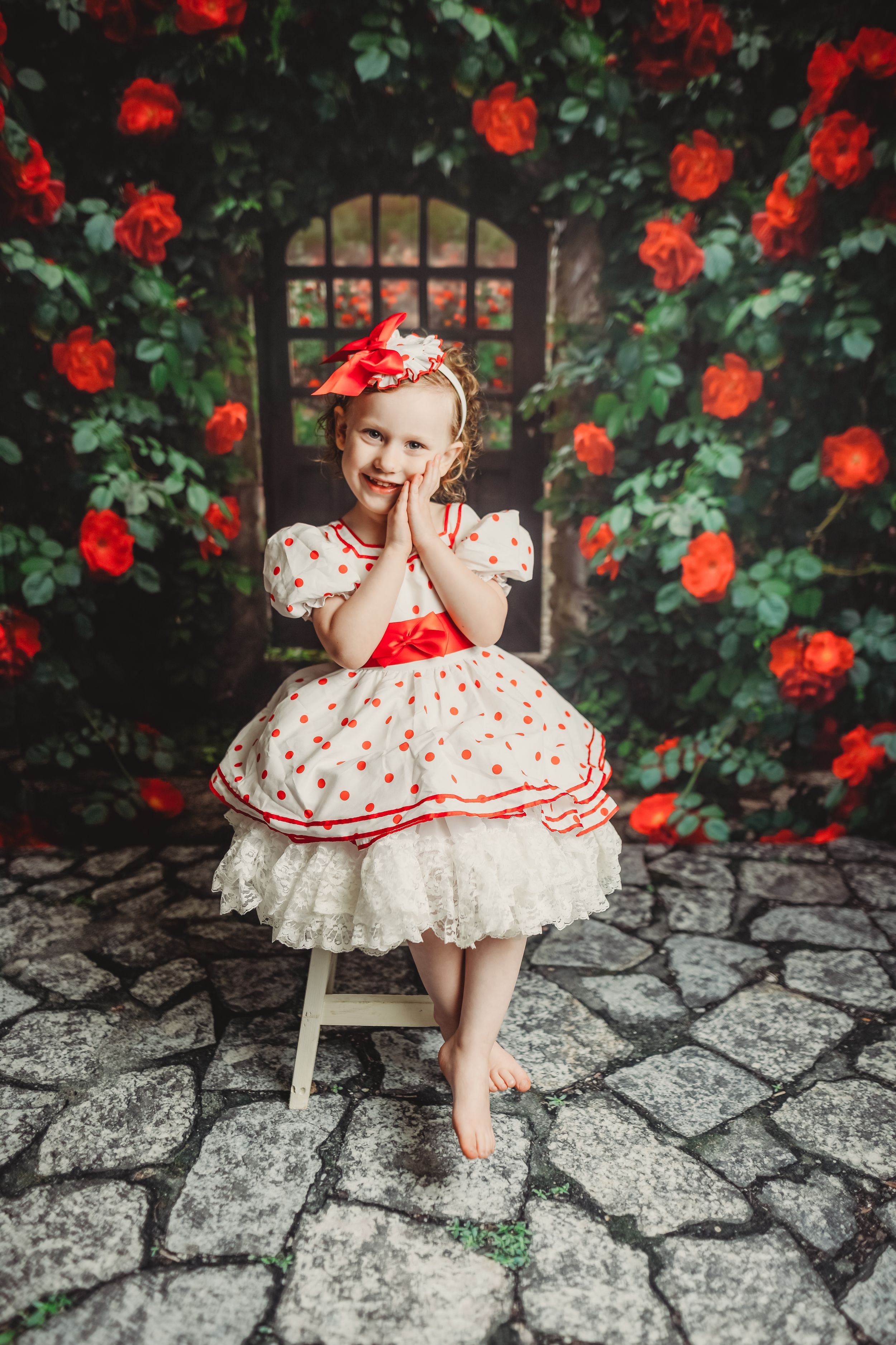 Young girl in white polka dot dress with red bow sits barefoot among blooming red roses in a garden.