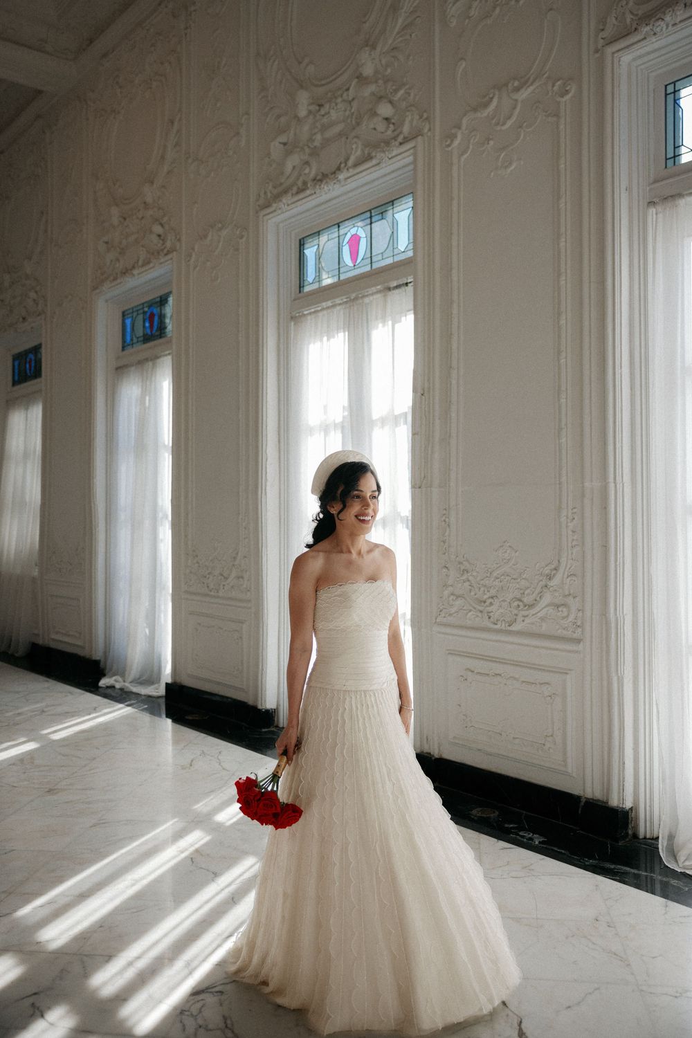 Bride in strapless white mermaid gown holding red bouquet in ornate white ballroom with marble floors.