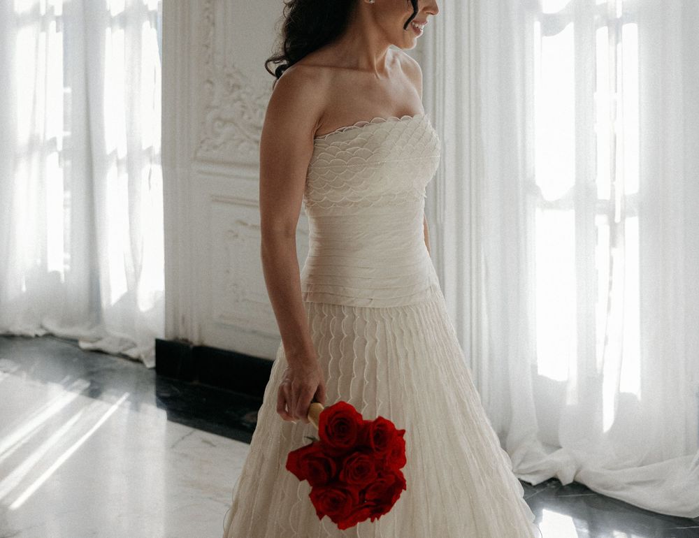Bride in strapless ivory wedding gown holding red rose bouquet standing by sheer white curtains in elegant room.