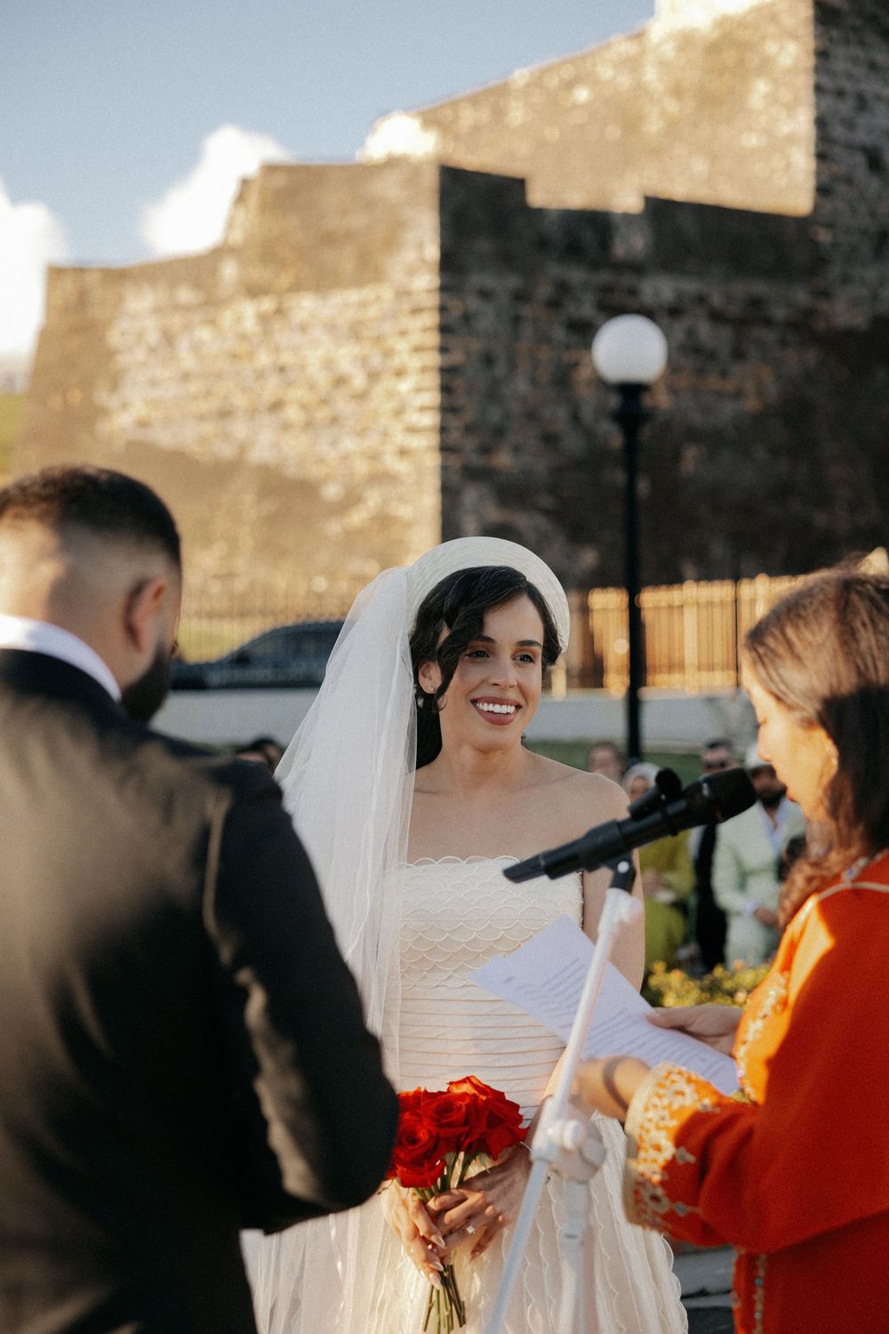 Smiling bride in white gown and veil holds red roses during outdoor wedding ceremony with officiant.