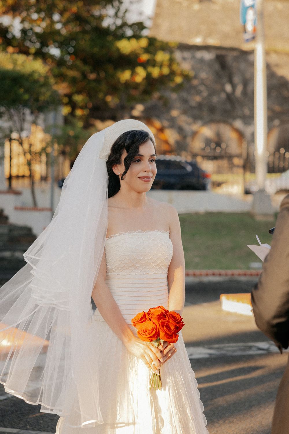 Bride in white strapless gown and veil holding vibrant orange rose bouquet, smiling outdoors on sunny wedding day.
