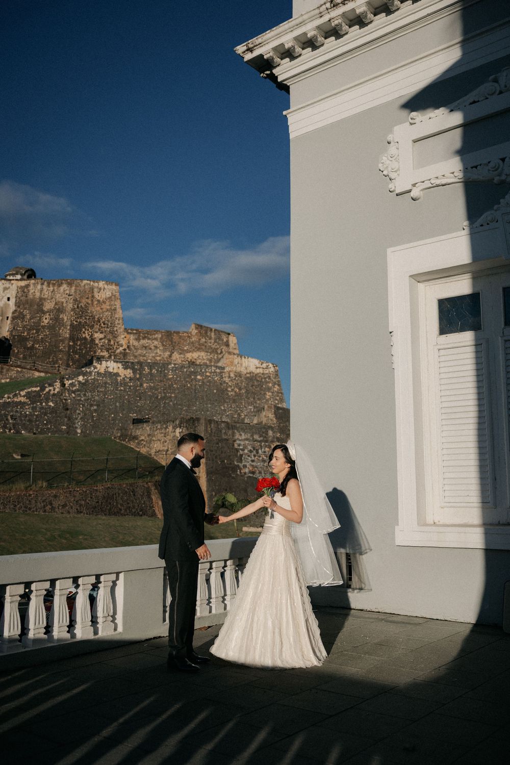 Bride and groom share a romantic moment on a balcony with a historic fortress backdrop in Puerto Rico.