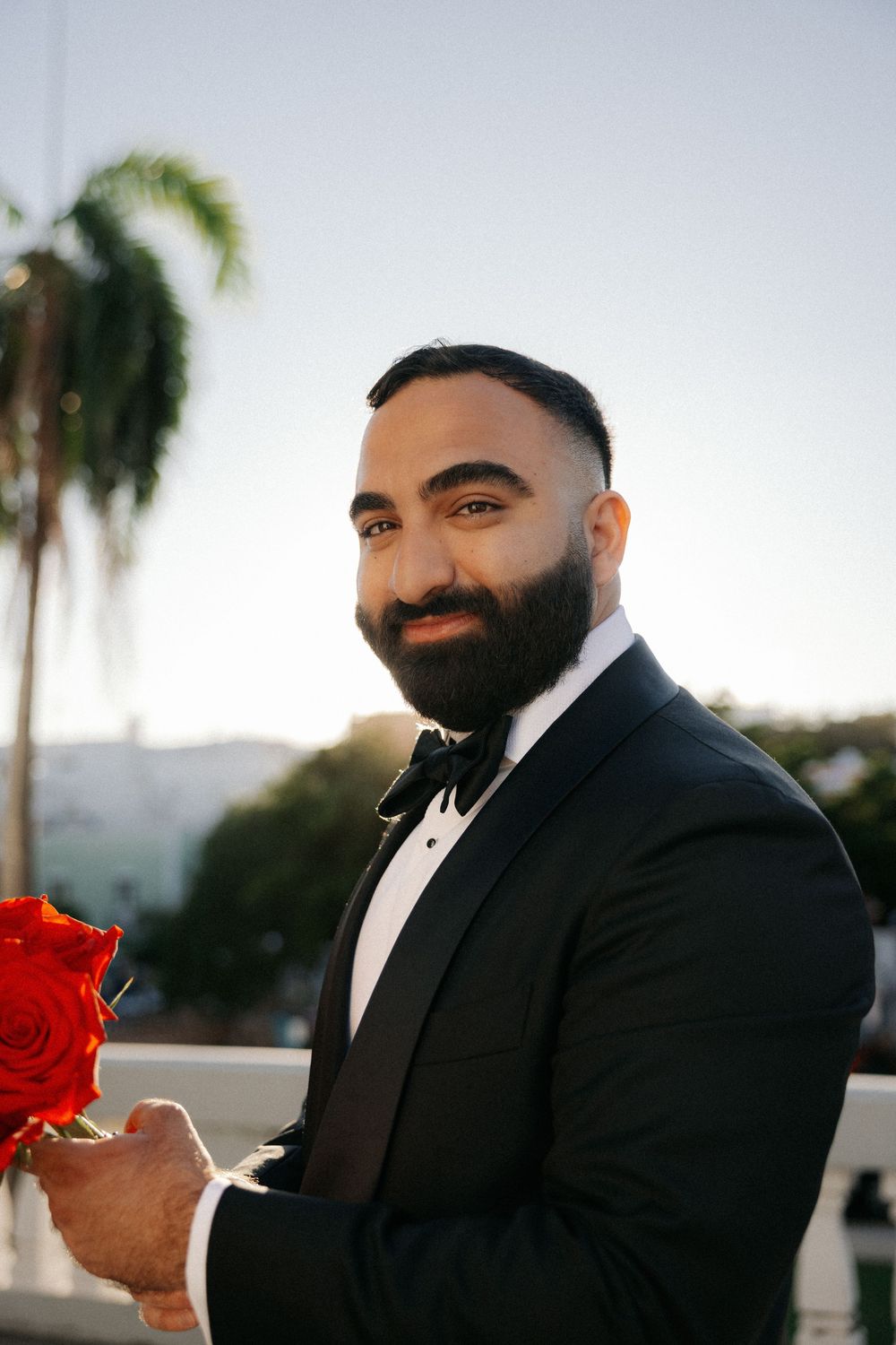 Bearded man in black tuxedo with bow tie holding red roses outdoors with palm trees at sunset.