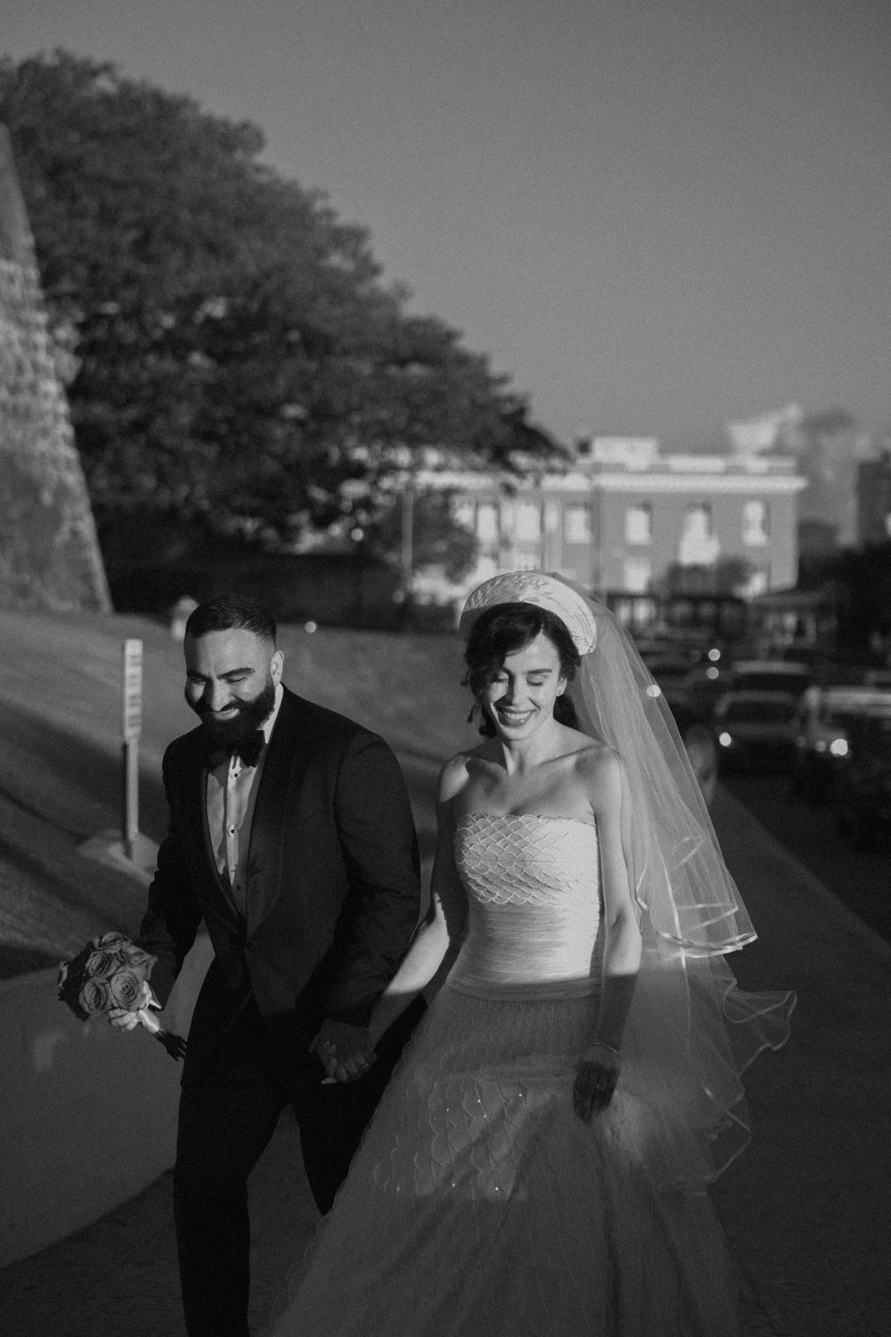 Black and white photo of a smiling bride in a veil and groom in a tuxedo walking together on a street.