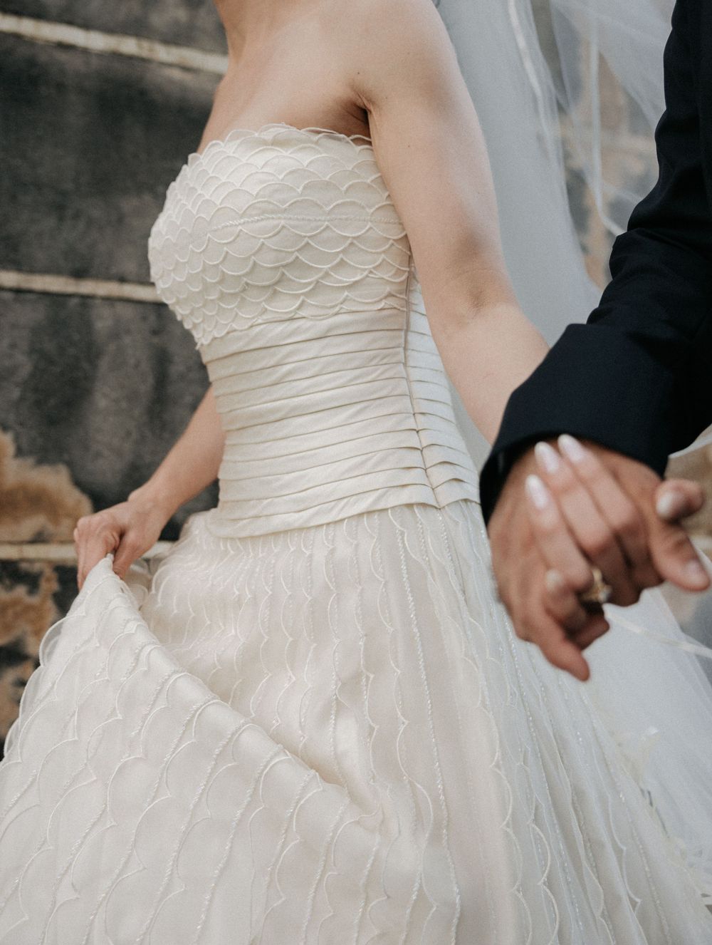 Bride in strapless white wedding gown with scalloped bodice holds groom's hand, veil flowing behind her.
