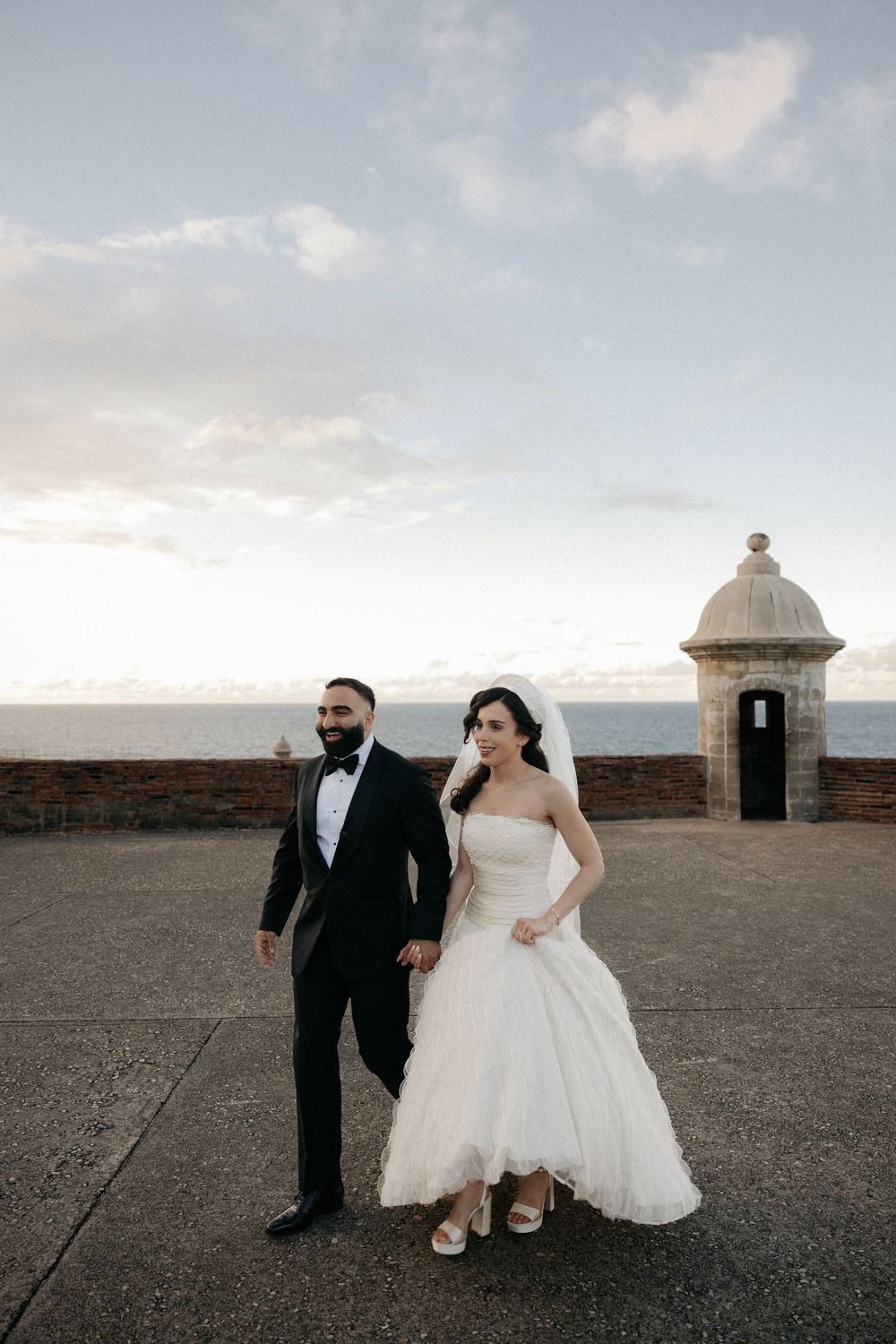 Bride and groom hold hands walking at El Morro fort in San Juan, Puerto Rico, with ocean views at sunset.