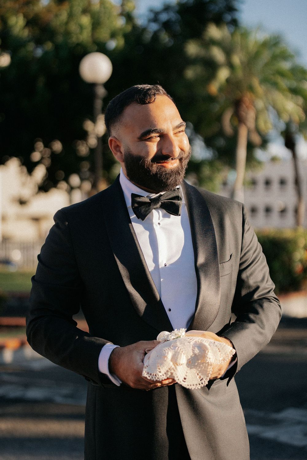 Smiling groom in black tuxedo and bow tie holds ring pillow outdoors with palm trees in background.
