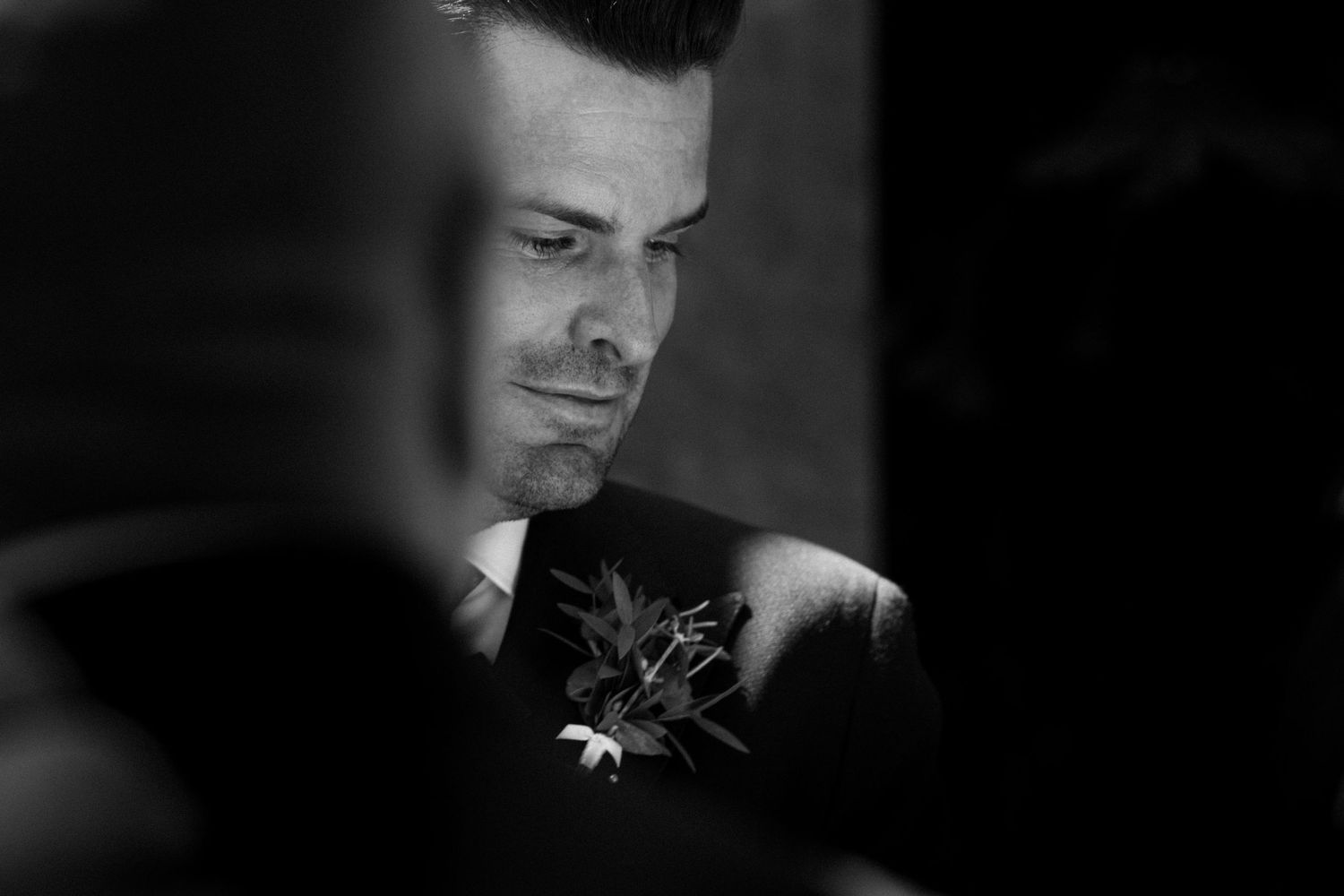 Black and white photo of a groom looking down at his boutonniere during wedding preparations.