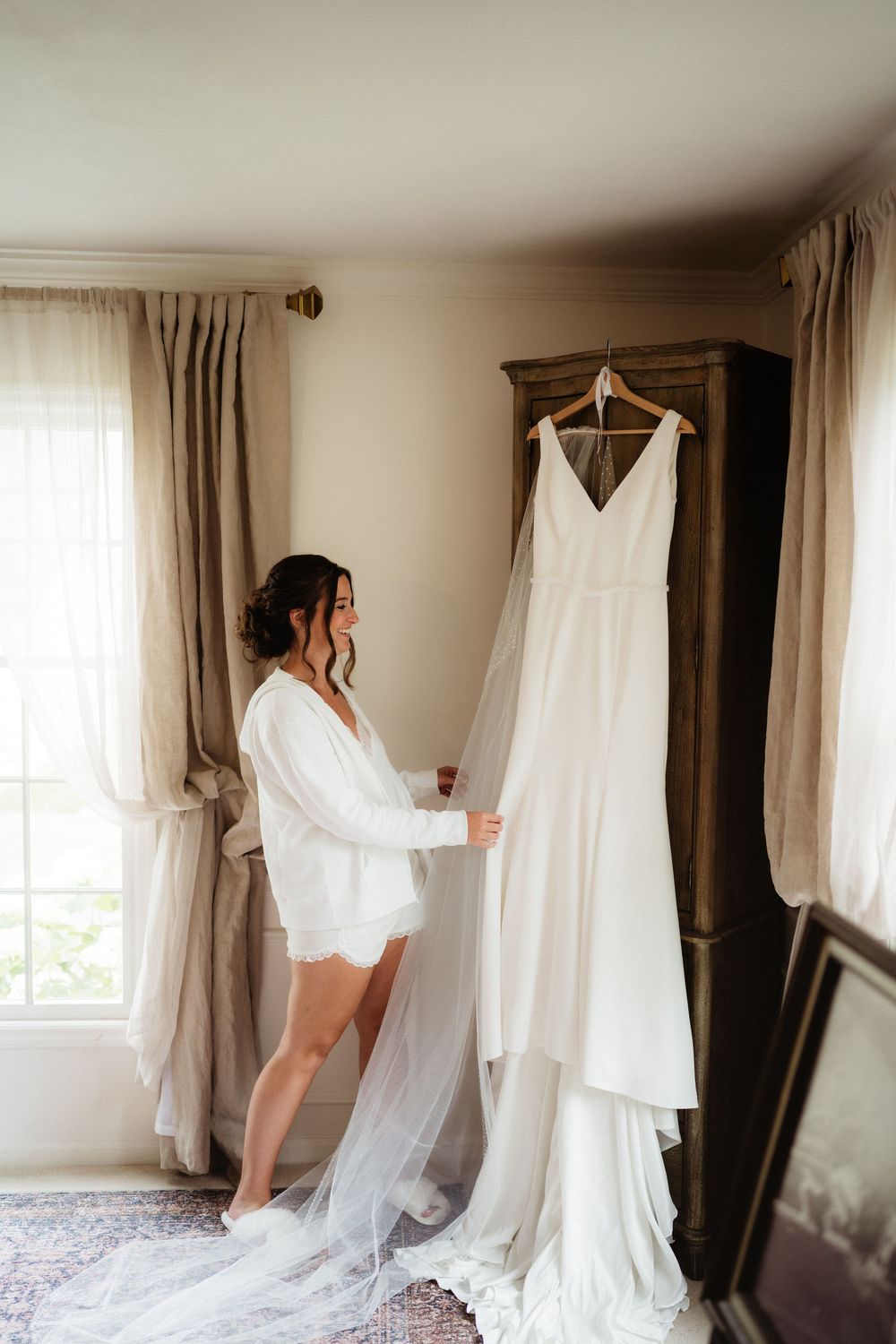 Bride in white robe admiring her elegant wedding gown hanging on a wooden wardrobe in a softly lit bridal suite.