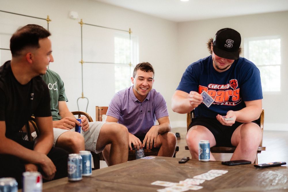 Three young men playing a card game at a wooden table with canned drinks, smiling and enjoying a casual indoor gathering.