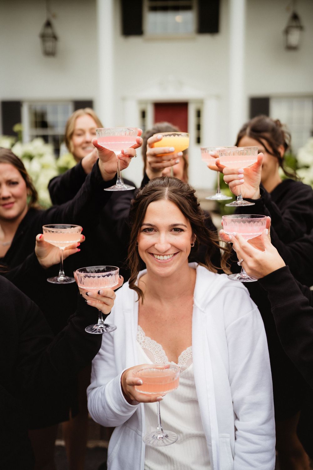 Bride in white robe smiles holding pink cocktail while bridesmaids in black robes raise glasses in a toast outside.