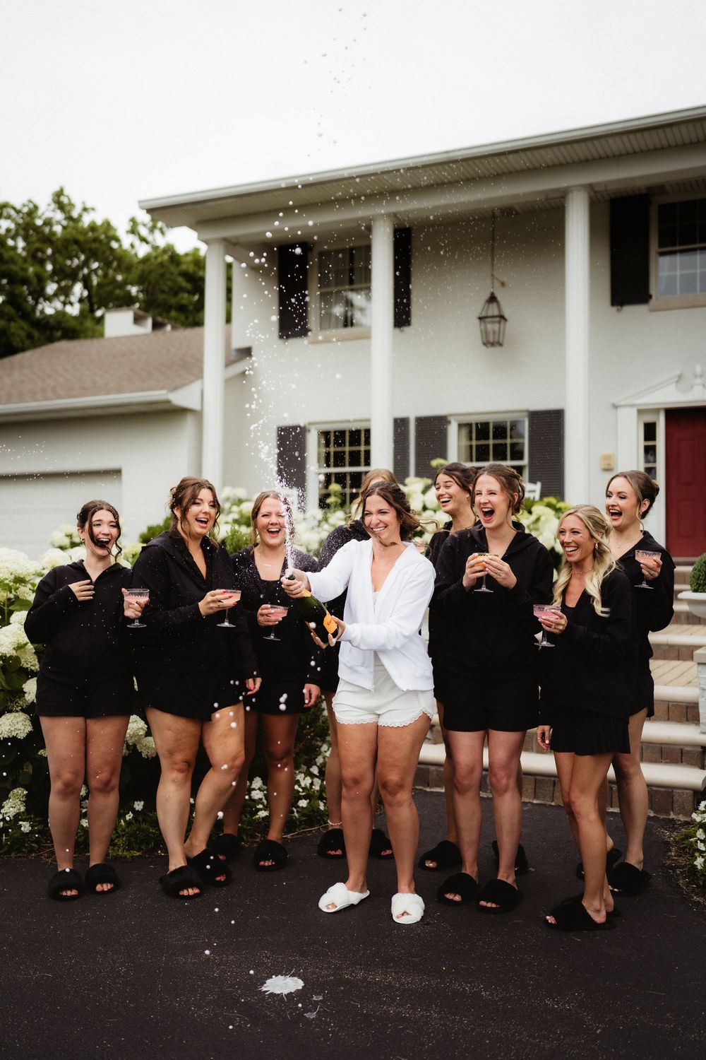 Bride in white pajamas pops champagne with bridesmaids in black robes outside a white colonial home.