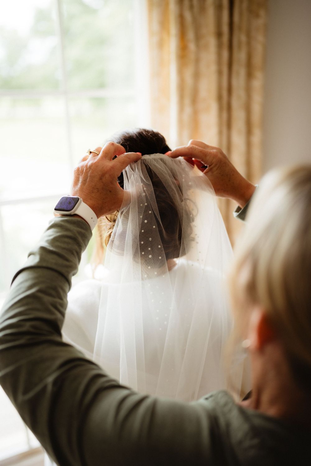 A woman helps a bride put on a delicate polka dot wedding veil near a bright window before the ceremony.