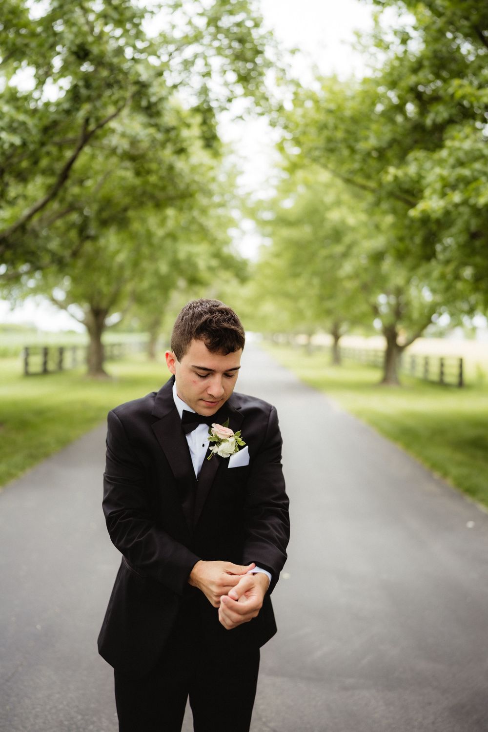 Groom in black tuxedo with white boutonniere stands on tree-lined path, looking down at his hands.