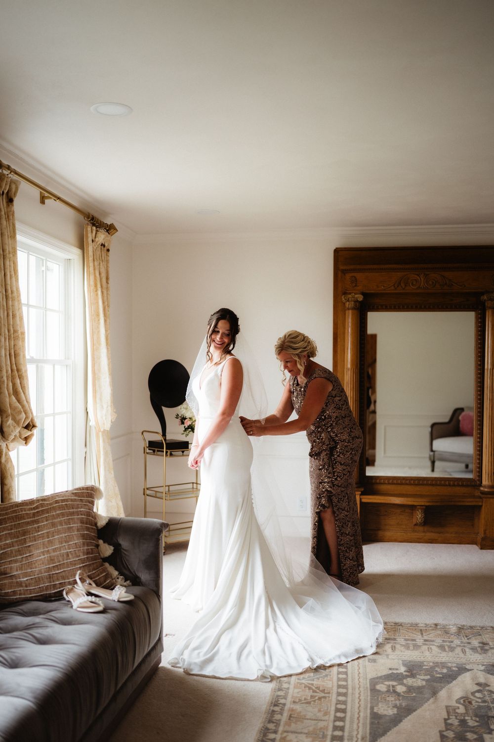 A bride in a white gown gets her dress fastened by a woman in a floral dress in an elegant, softly lit room.