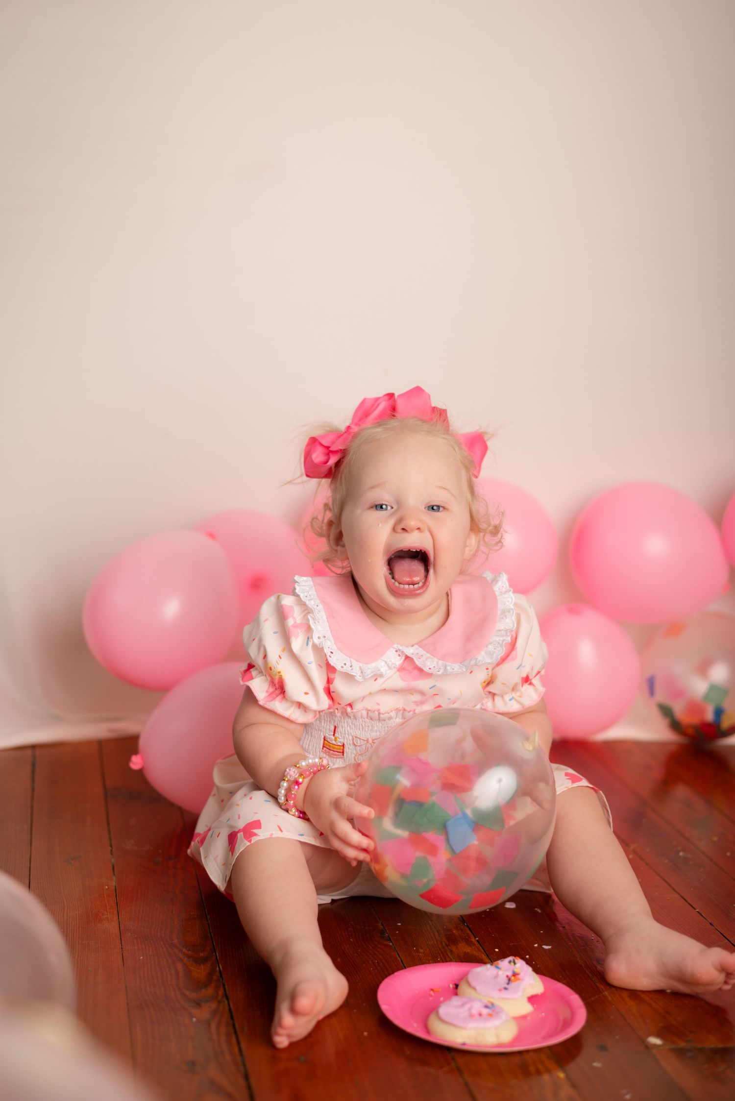 Baby girl in pink dress holds candy-filled balloon, surrounded by pink balloons, celebrating her first birthday.