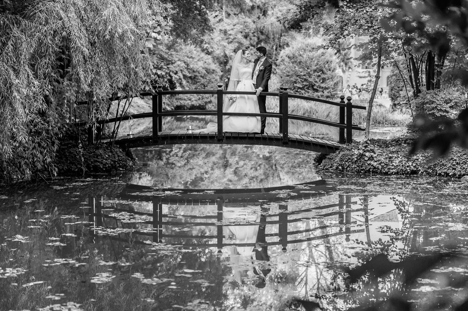 Black and white photo of couple kissing on an arched wooden bridge reflected in calm pond water surrounded by lush trees.