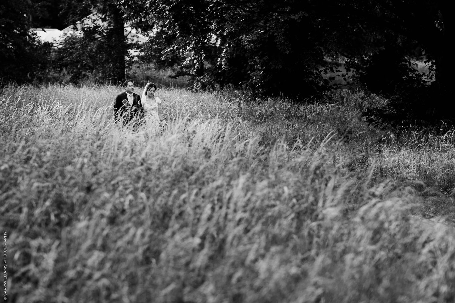 Black and white photo of a couple walking through tall grass in a field with trees in the background.