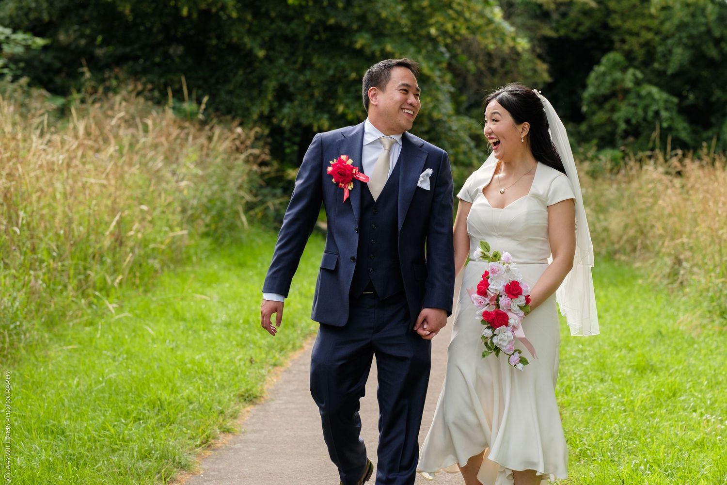 A happy bride and groom walk hand-in-hand along a garden path, she in a white dress and veil, he in a navy suit.