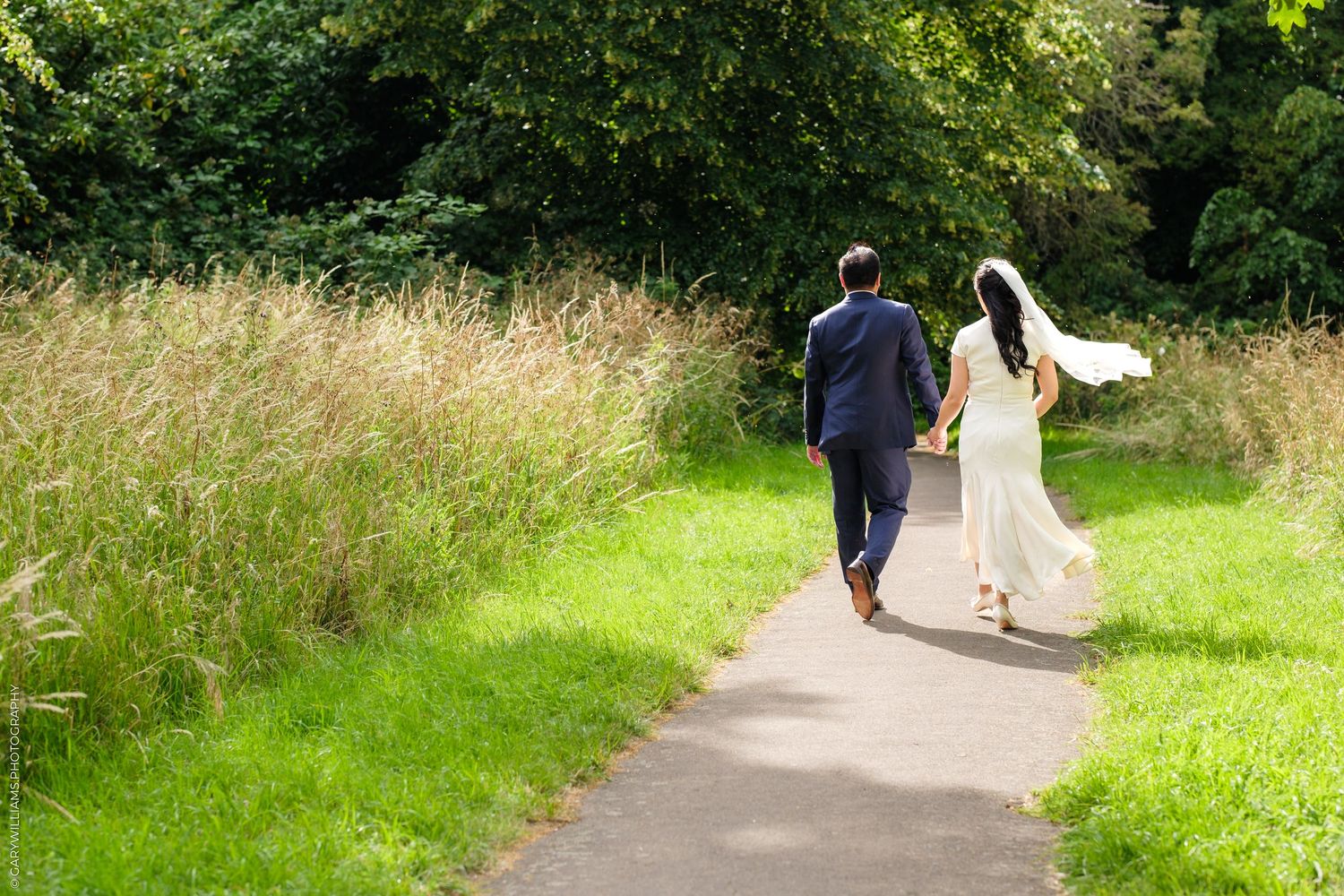 Newlywed couple walking hand in hand down a garden path, bride in white gown with veil flowing in the breeze.