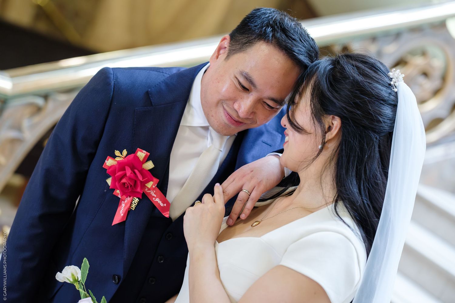 A couple in wedding attire share a romantic moment, groom in navy suit with red boutonniere, bride in white dress and veil.