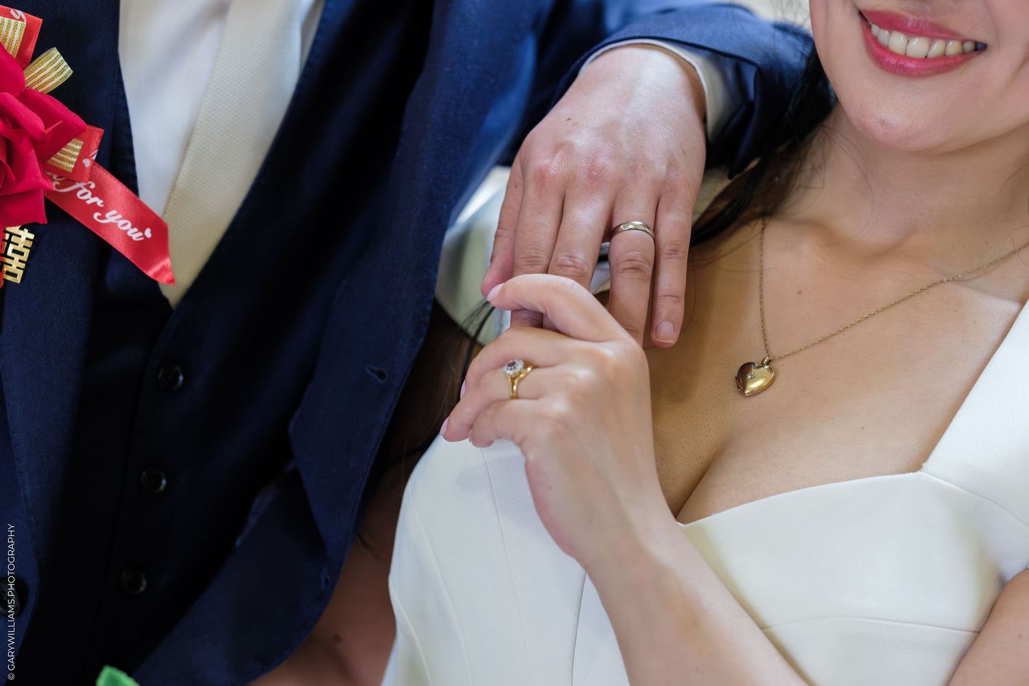 Close-up of bride and groom holding hands, showing wedding rings, bride in white dress with gold necklace.