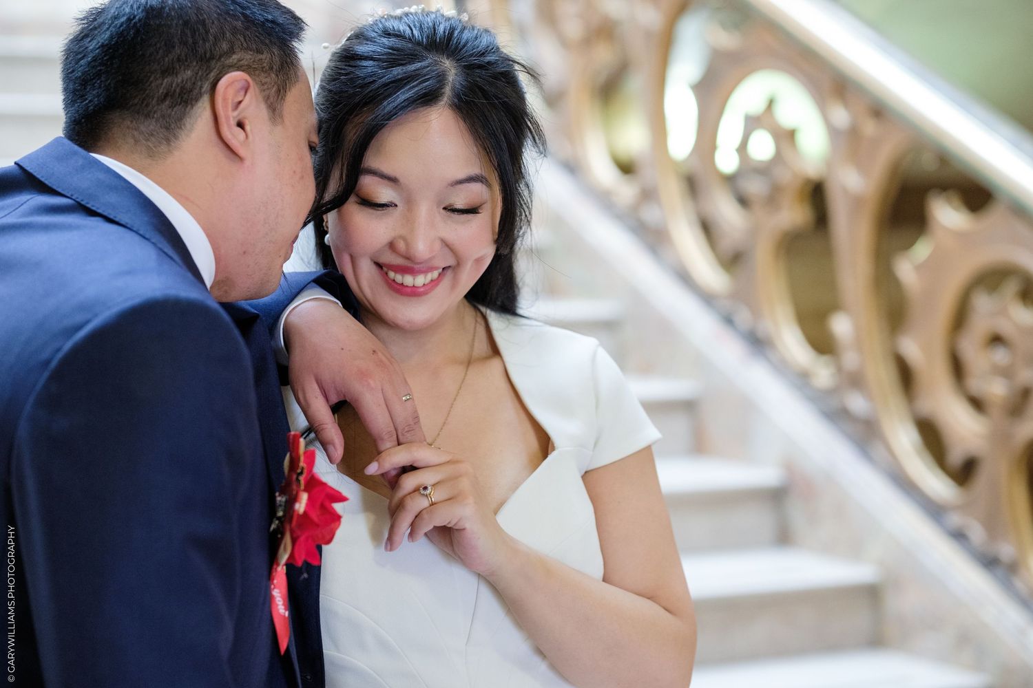 Smiling bride in white dress holds groom's arm on ornate staircase during elegant wedding ceremony.