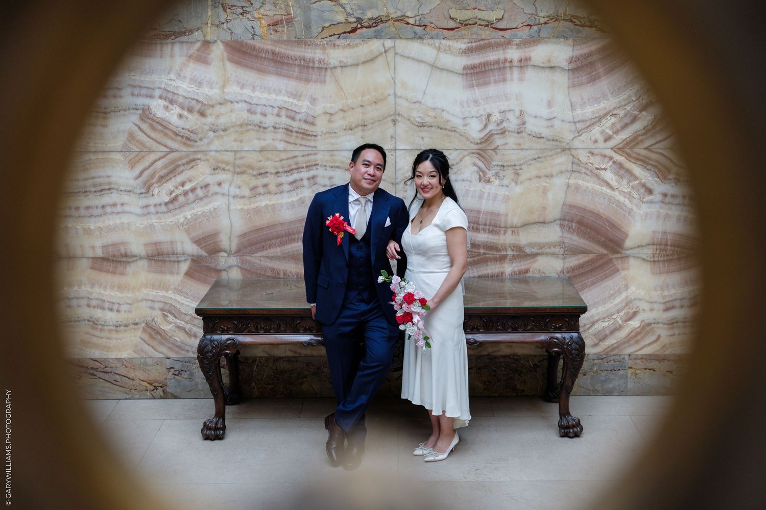 Newlywed couple posing together in elegant marble hall, groom in navy suit, bride in white dress holding red roses.