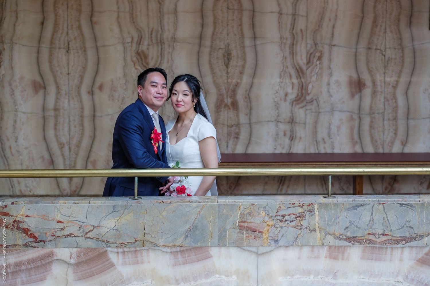 Asian newlywed couple posing on balcony in wedding attire.