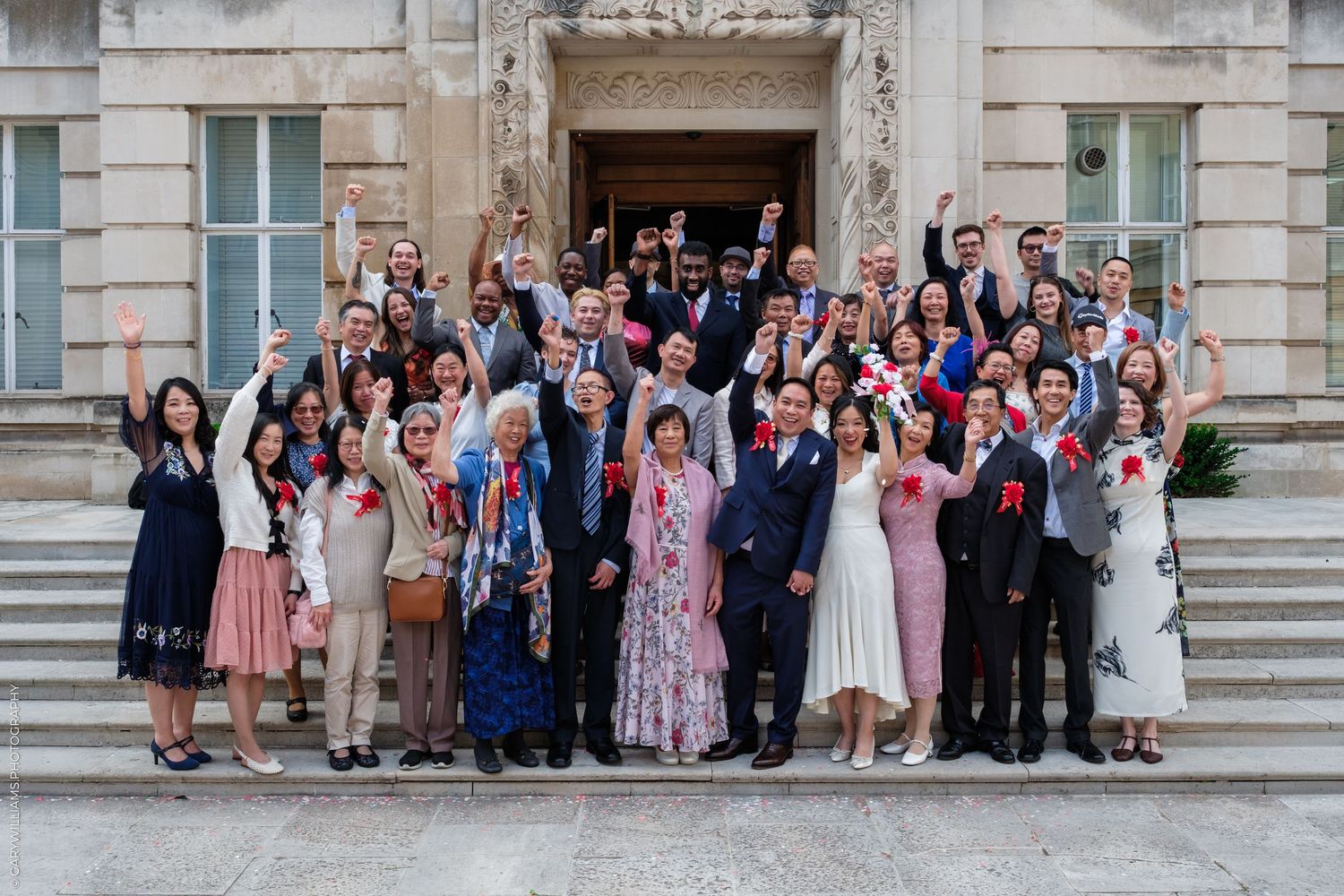 Large group of elegantly dressed people celebrating on stone steps outside a grand building, raising hands joyfully.