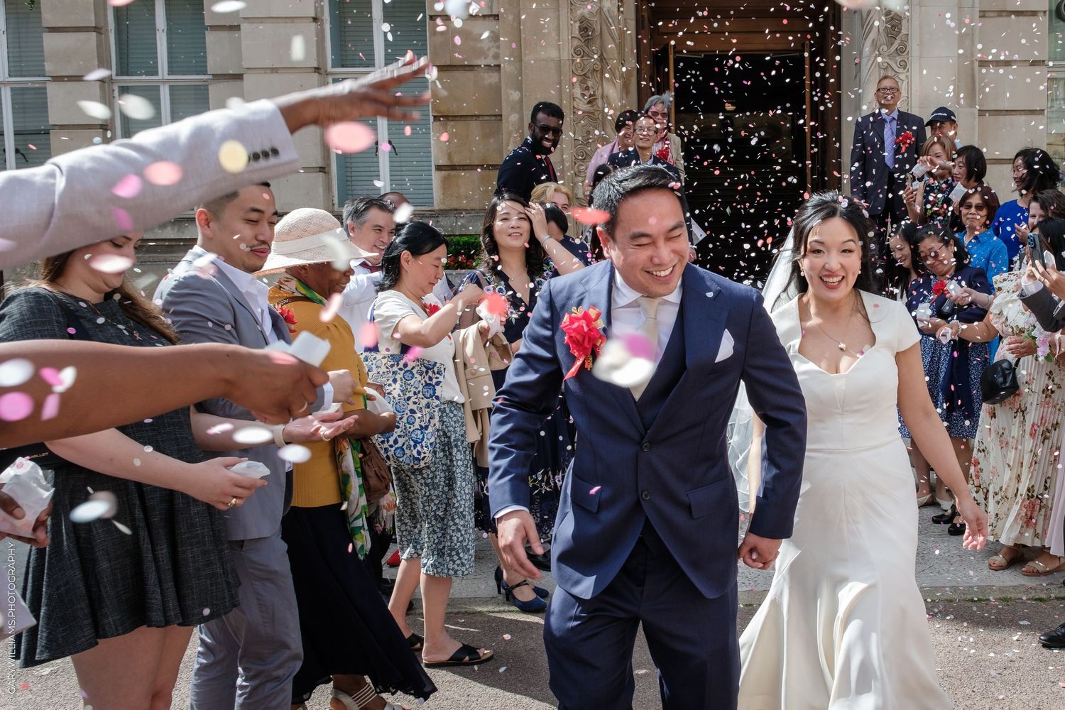 Newlywed couple walking through confetti shower surrounded by cheering wedding guests outdoors on sunny day.