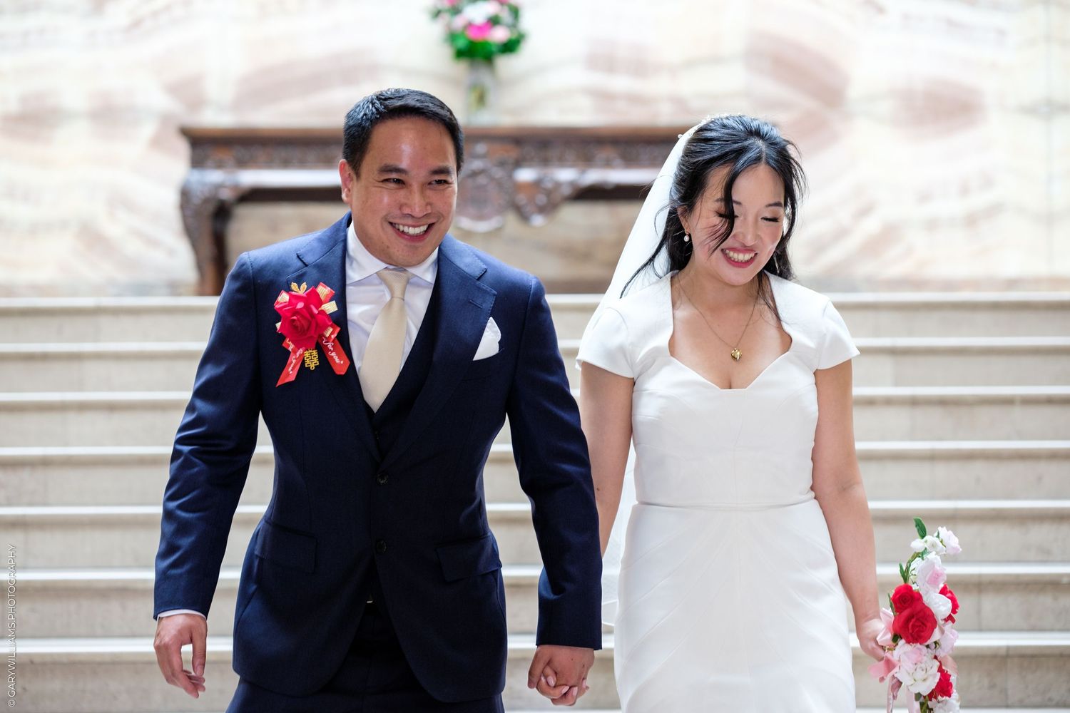 Smiling bride and groom walking hand in hand down stairs, bride holding pink bouquet in wedding venue.
