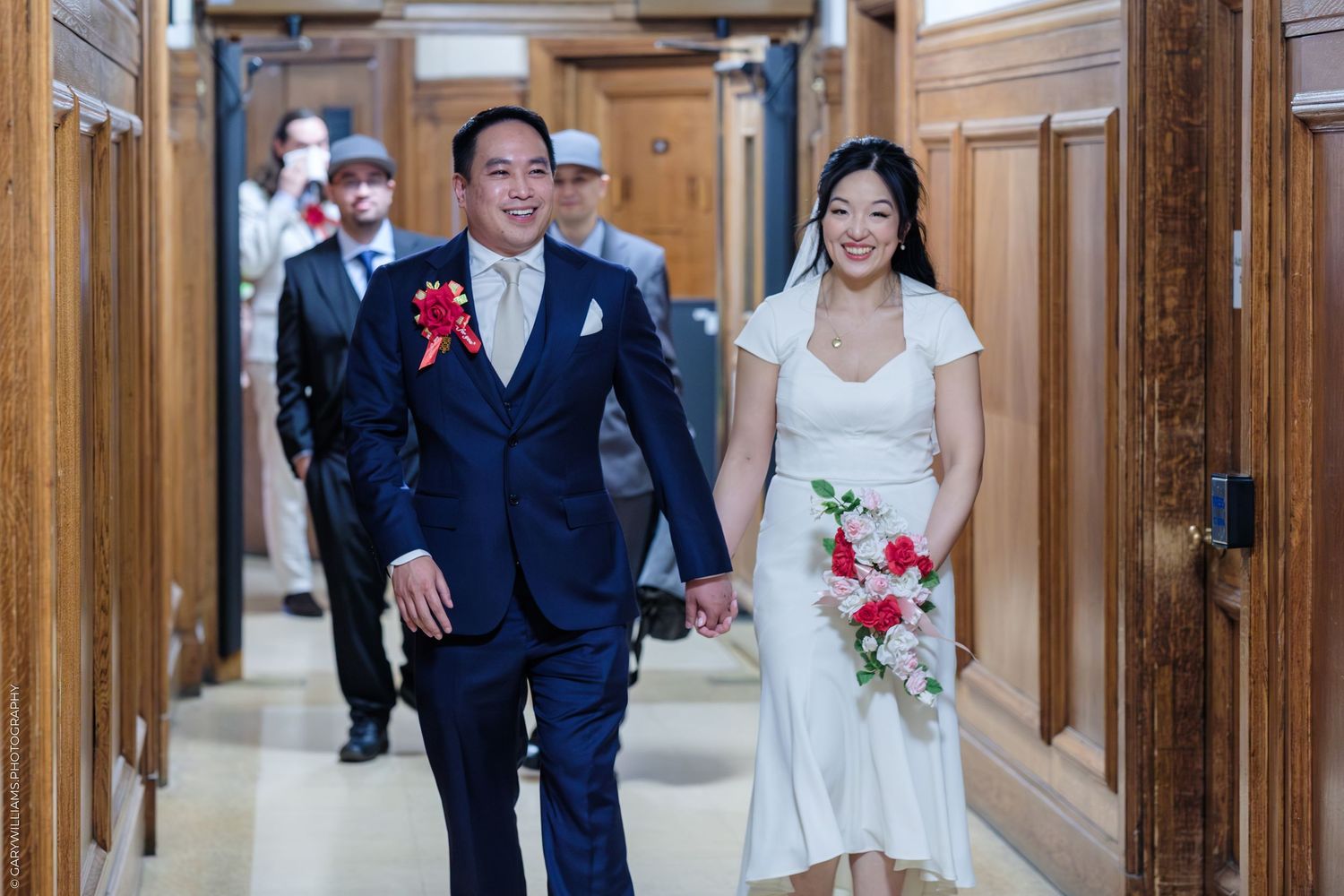 Smiling newlywed couple walks hand-in-hand down a hallway, groom in navy suit with red boutonniere, bride in white dress.