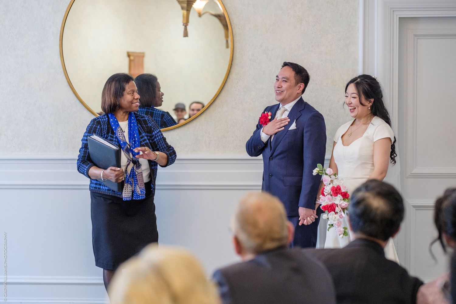 Officiant smiling with Asian bride and groom during intimate wedding ceremony in elegant white room.