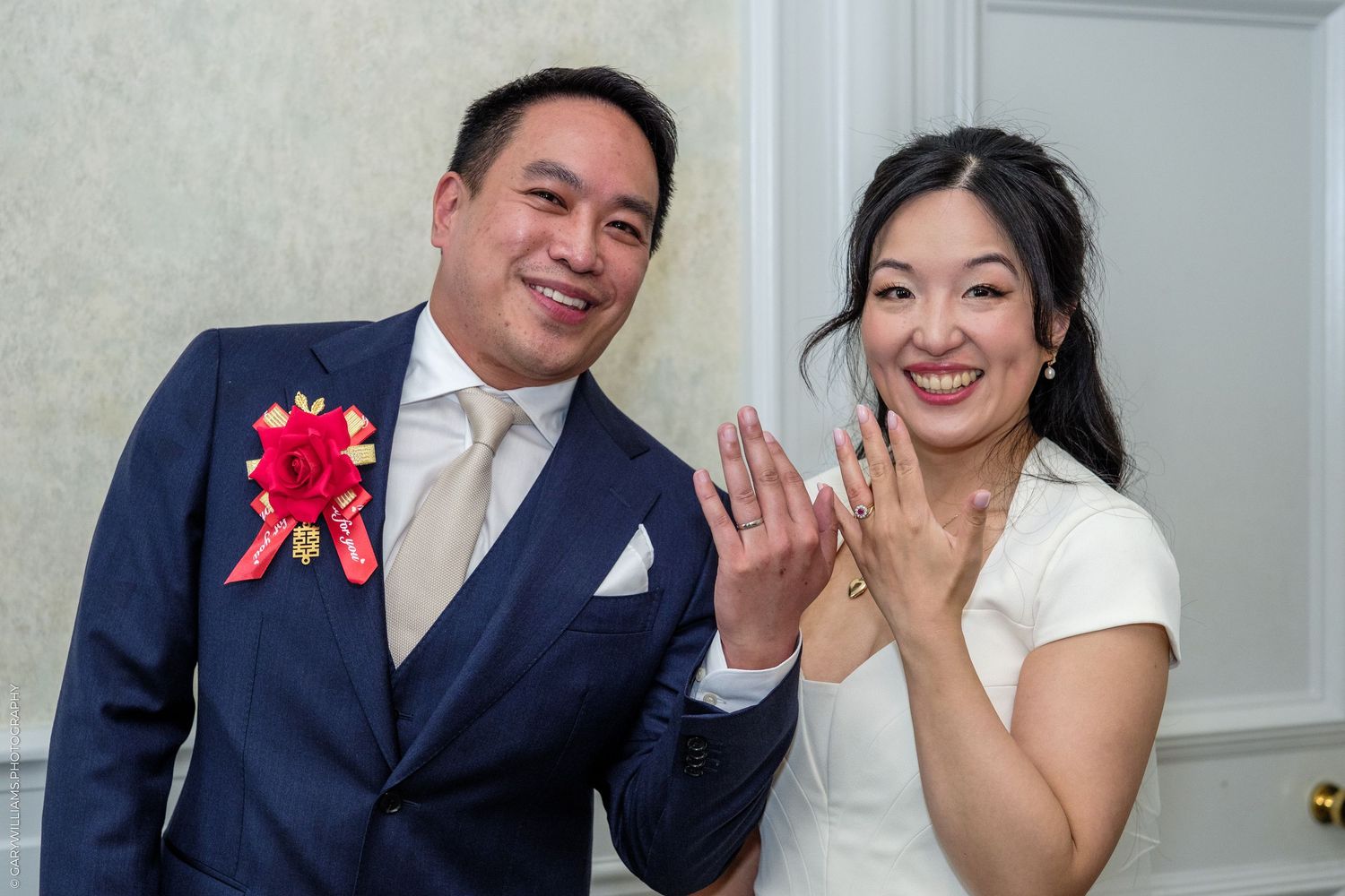 Newlywed Asian couple smiling and showing wedding rings, groom wearing navy suit with red floral boutonniere indoors.